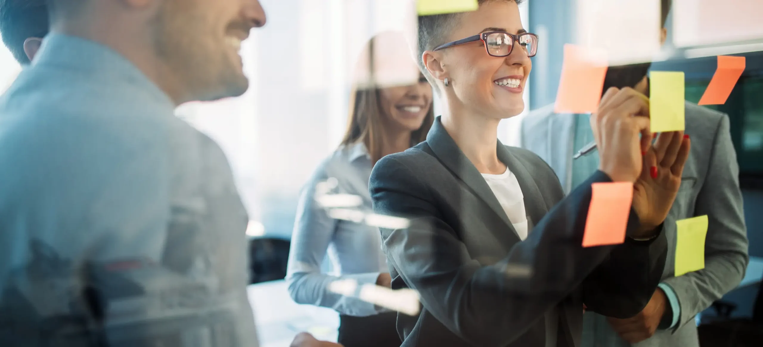 A diverse group of smiling professionals collaborating in a bright office. A woman with glasses is writing on a glass wall covered in colorful sticky notes, representing teamwork and strategic planning.