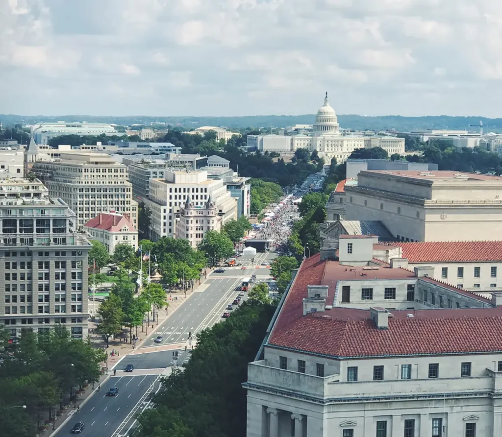 A high-angle, wide-view landscape of Washington D.C., showing a long, tree-lined avenue leading directly to the United States Capitol Building under a partly cloudy sky.