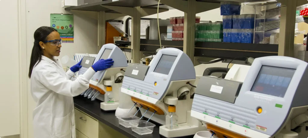 A laboratory professional in a white coat, safety glasses, and blue gloves operating a row of automated medical diagnostic machines in a clean lab setting.