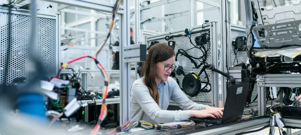 A female technician wearing glasses and a lab coat working on a laptop at a high-tech workstation filled with complex wiring and electronic components.
