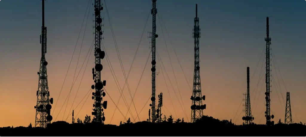 A row of tall telecommunication towers and antennas silhouetted against a gradient sunset sky of orange and blue.