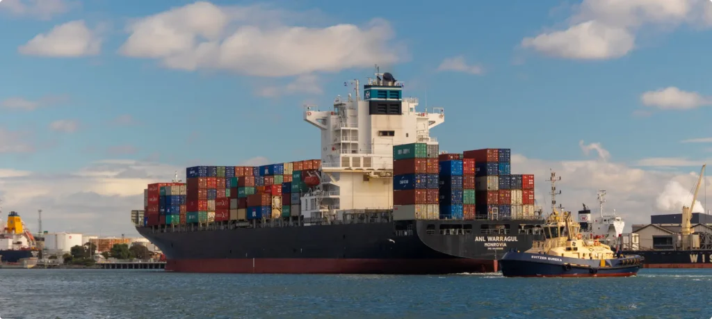A massive container ship, the "ANL Warragul," loaded with colorful shipping containers, being guided through a harbor by a small tugboat.