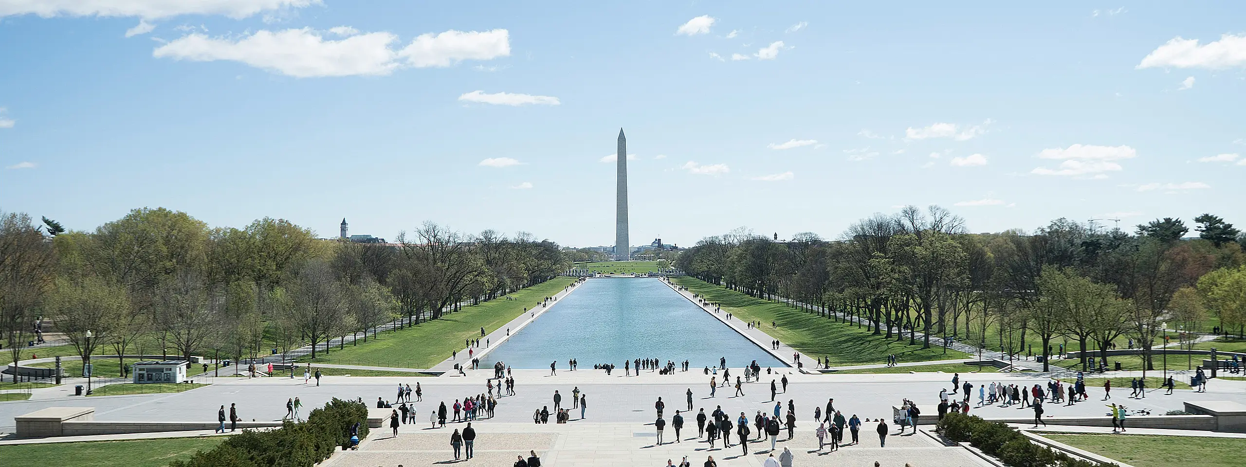 A wide, high-angle view of the National Mall in Washington, D.C. The Lincoln Memorial Reflecting Pool stretches toward the Washington Monument under a clear blue sky, with many people walking along the surrounding paths and green lawns.