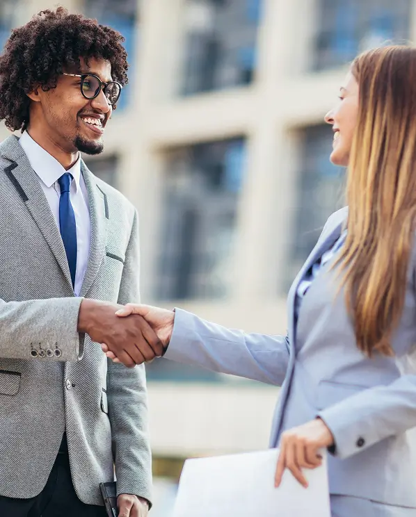 A man and woman in business suits smiling and shaking hands outdoors in front of a blurred office building.