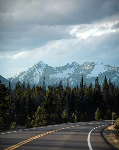A winding two-lane highway leading toward snow-capped mountain peaks under a dramatic, cloudy sky.